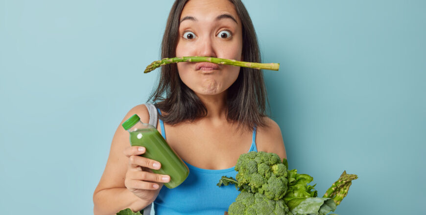 Boost your immune system. Surprised funny young Asian woman pouts lips with asparagus uses fresh green vegetables for making detox beverage keeps to healthy diet poses with homemade smoothie