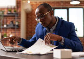 Business man analyzing papers on clipboard to plan marketing strategy with laptop. Entrepreneur using computer and financial documents to work on project planning in startup office.