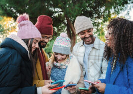 Group of multiracial young friends with coats and hats, smiling and watching the social media with a cellphone app. Portrait of happy people having fun sharing media and laughing with a smart phone. Communication concept. High quality photo