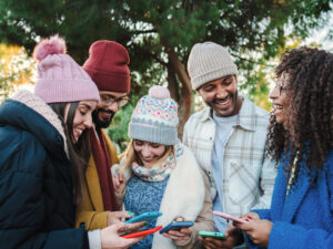 Group of multiracial young friends with coats and hats, smiling and watching the social media with a cellphone app. Portrait of happy people having fun sharing media and laughing with a smart phone. Communication concept. High quality photo