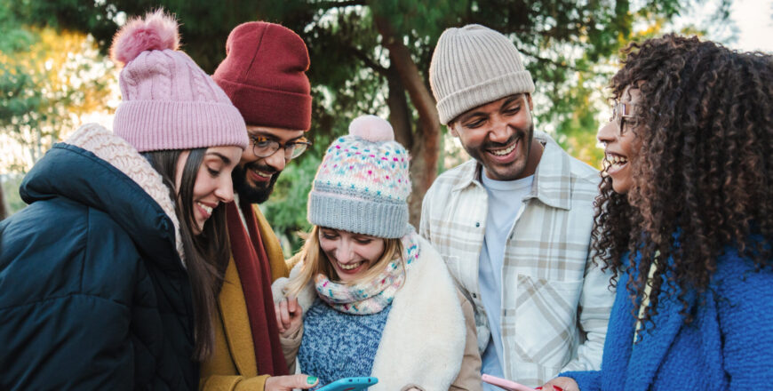 Group of multiracial young friends with coats and hats, smiling and watching the social media with a cellphone app. Portrait of happy people having fun sharing media and laughing with a smart phone. Communication concept