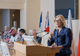 Mature female speaker in elegant suit making report while standing by tribune against row of foreign colleagues