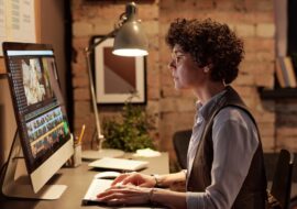 Young professional colorist sitting at her workplace in front of computer monitor and editing photos on special software