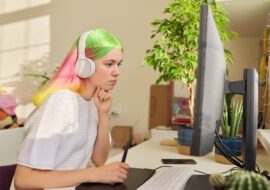 Girl teenager artist in headphones sitting at the table at home drawing on computer using graphics tablet. Hobbies and leisure of creative college student