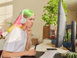 Girl teenager artist in headphones sitting at the table at home drawing on computer using graphics tablet. Hobbies and leisure of creative college student